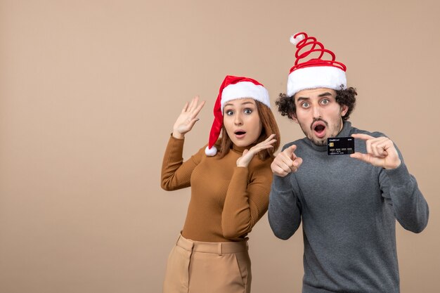 銀行カードを示す赤いサンタクロースの帽子の男を身に着けている興奮した満足のいくクールなカップルとクリスマス気分