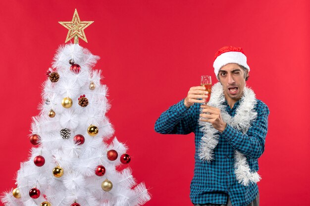 xmas mood with funny young man with santa claus hat in a blue stripped shirt raising a glass of wine near Christmas tree