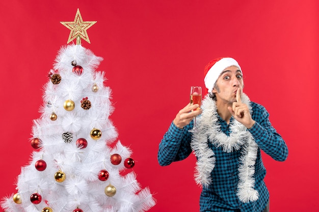 Umore natalizio con felice pazzo emotivo giovane uomo con cappello di babbo natale in una camicia blu spogliata alzando un bicchiere di vino facendo gesto di silenzio vicino all'albero di natale