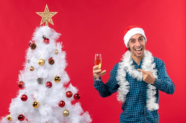 Umore natalizio con felice giovane uomo con cappello di babbo natale in una camicia blu spogliata che tiene un bicchiere di vino vicino all'albero di natale