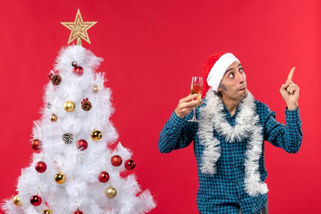 Atmosfera natalizia con felice giovane uomo con cappello di babbo natale in una maglietta spogliata blu alzando un bicchiere di vino che mostra quanto sopra vicino all'albero di natale