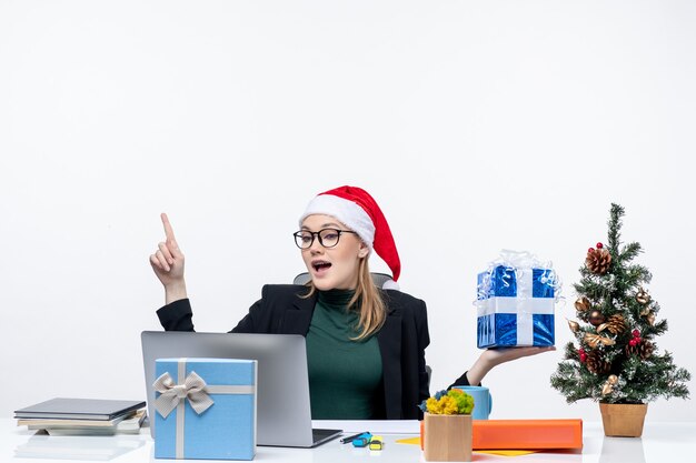 Рождественское настроение с позитивной взволнованной забавной молодой женщиной в шляпе санта-клауса и в очках, сидящей за столом, показывающим подарок, указывающим выше на белом фоне