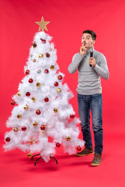 xmas mood with positive guy dressed in jeans standing near decorated Christmas tree and holding microphone and making silence gesture