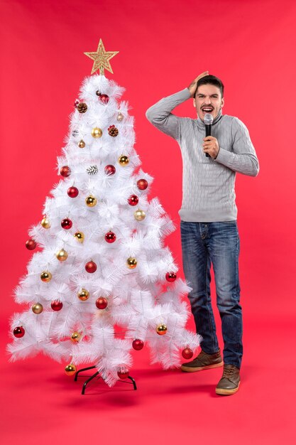 новогоднее настроение с позитивным парнем в джинсах, стоящим возле украшенной елки, с микрофоном и рукой в голове