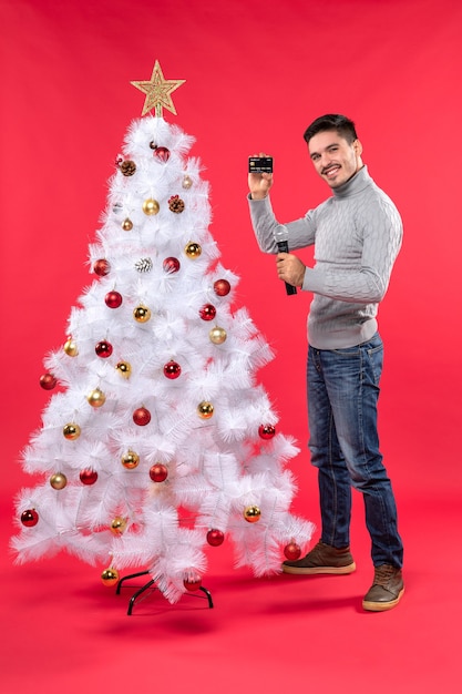 xmas mood with serious young guy standing near decorated Christmas tree and holding microphone taking photo