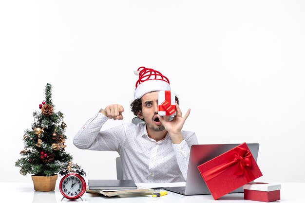 彼の顔に彼の贈り物を上げ、白い背景に何かを指しているサンタクロースの帽子をかぶったショックを受けた興奮したビジネスパーソンとクリスマス気分