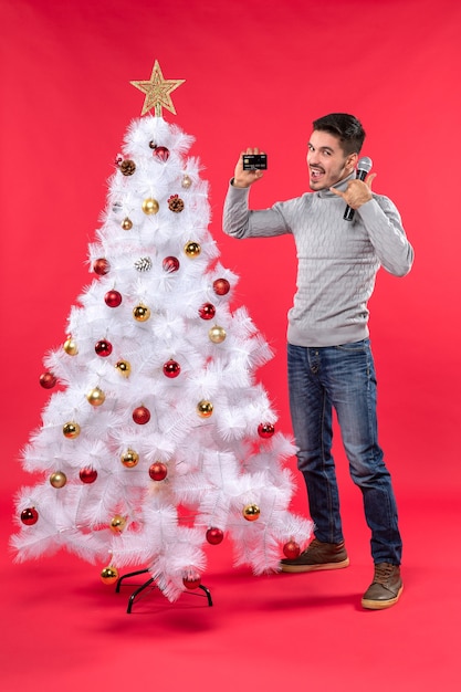 xmas mood with smiling young guy standing near decorated Christmas tree and holding microphone and phone making call me gesture