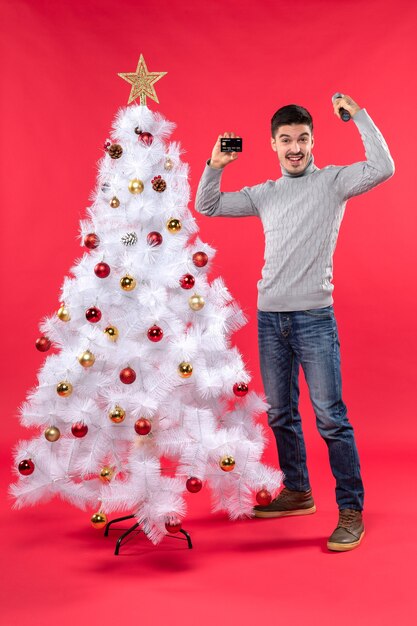 xmas mood with smiling young guy standing near decorated Christmas tree and holding microphone and phone showing his power