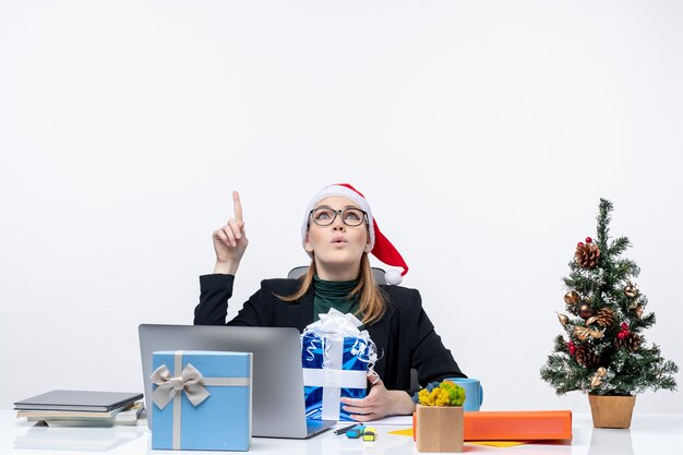 Рождественское настроение с удивленной молодой женщиной в шляпе санта-клауса и очках, сидящей за столом с подарком и показывающим выше на белом фоне
