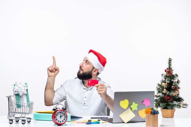 Free photo xmas mood with young adult with santa claus hat and holding bank card and looking pointing above in the office