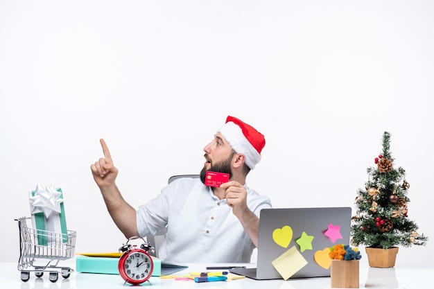 サンタクロースの帽子をかぶって銀行カードを持って、オフィスで意外にも左上を指している若い大人とのクリスマス気分