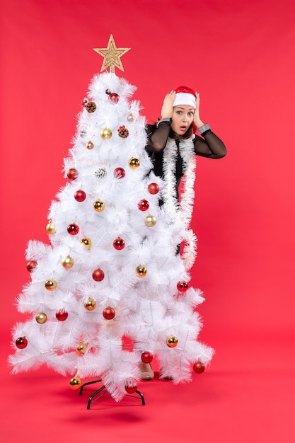 Рождественское настроение с молодой красивой девушкой в черном платье с шляпой санта-клауса, прячущейся за новогодней елкой в глубоких мыслях о красном