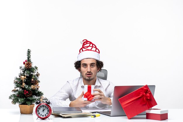 オフィスに座って、白い背景の上のカメラにポーズをとって彼の贈り物を保持しているサンタクロースの帽子をかぶった青年実業家とクリスマス気分