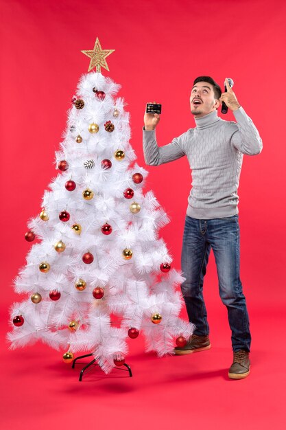 xmas mood with young guy standing near decorated Christmas tree and holding microphone and phone lookin above