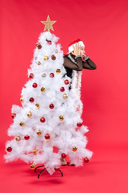 Рождественское настроение с молодой потрясенной красивой девушкой в черном платье с шапкой санта-клауса, прячущейся за новогодней елкой