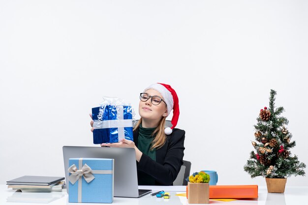 Xmas mood with young woman with santa claus hat and wearing eyeglasses sitting at a table looking at her gift proudly on white background