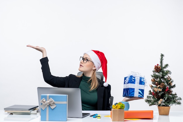 Atmosfera natalizia con giovane donna con cappello di babbo natale e occhiali da vista seduto a un tavolo che mostra il regalo che punta qualcosa su sfondo bianco