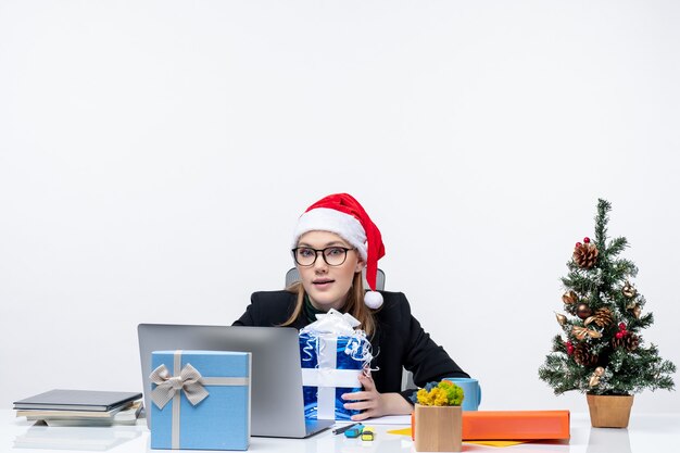 Рождественское настроение с молодой женщиной в шляпе санта-клауса и очках, сидящей за столом, где подарки и украшенная новогодняя елка на нем на белом фоне
