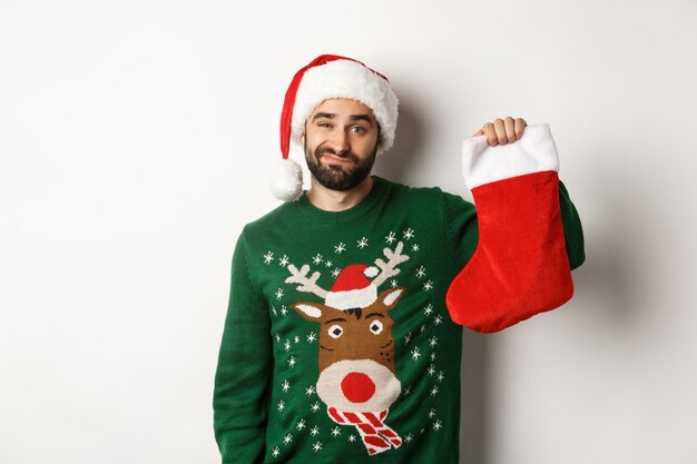 Xmas and winter holidays concept. Displeased guy disappointed in gift in Christmas sock, standing upset in Santa hat against white background