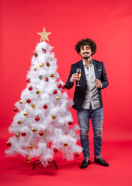 Xsmas celebration with bearded young man with wine standing near Christmas tree on red