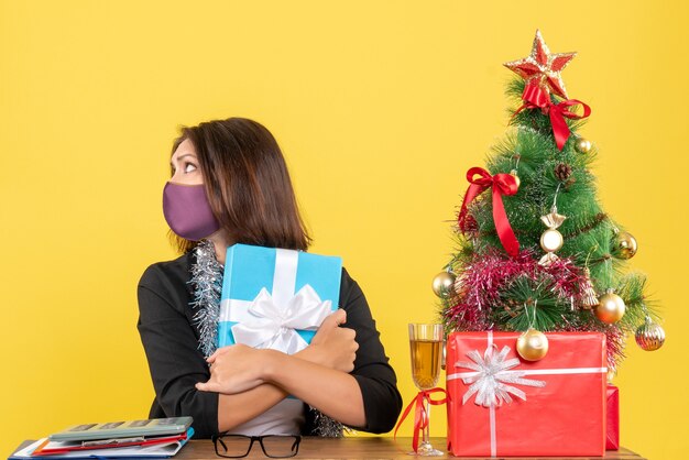 Xsmas mood with beautiful lady in suit with medical mask and embracing gift in the office on yellow 