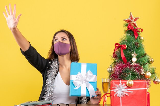 Xsmas mood with beautiful lady in suit with medical mask and holding gift saying hello in the office on yellow 