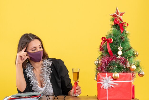 Xsmas mood with beautiful lady in suit with medical mask and raising wine in the office on yellow 