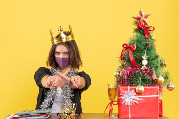 Рождественское настроение с красивой дамой в костюме с медицинской маской и маской, скрещивающей пальцы в офисе на желтом