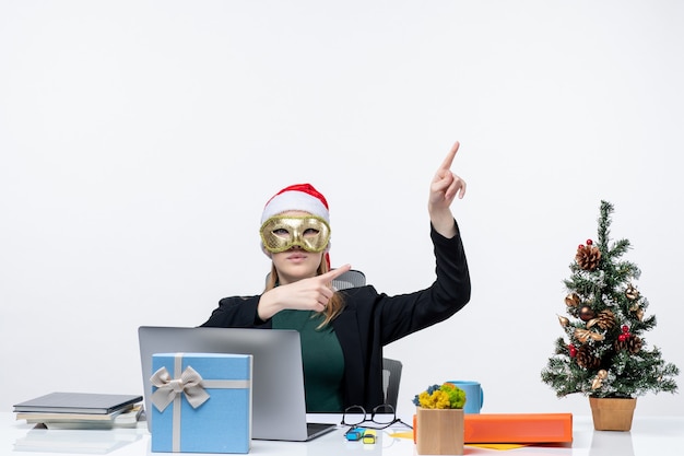 Xsmas mood with blonde young woman with santa claus hat and wearing mask sitting at a table pointing above on the left side on white background