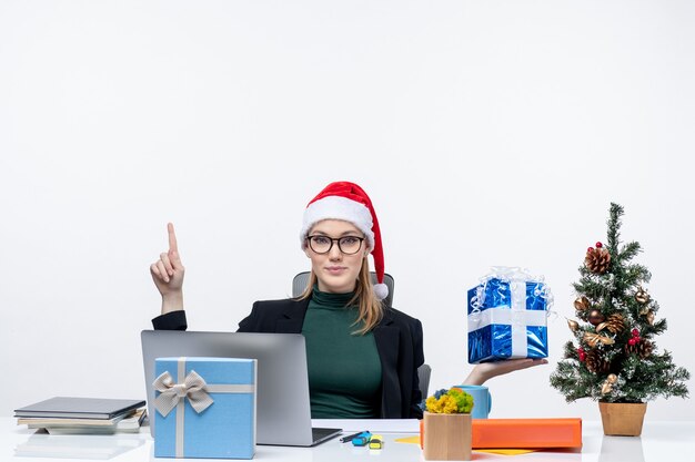 Xsmas mood with confident and serious young woman with santa claus hat and wearing eyeglasses sitting at a table showing gift pointing above on white background