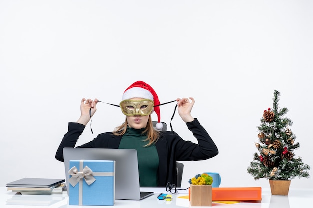 Atmosfera natalizia con curiosa giovane donna con cappello di babbo natale e maschera da portare seduto a un tavolo su sfondo bianco