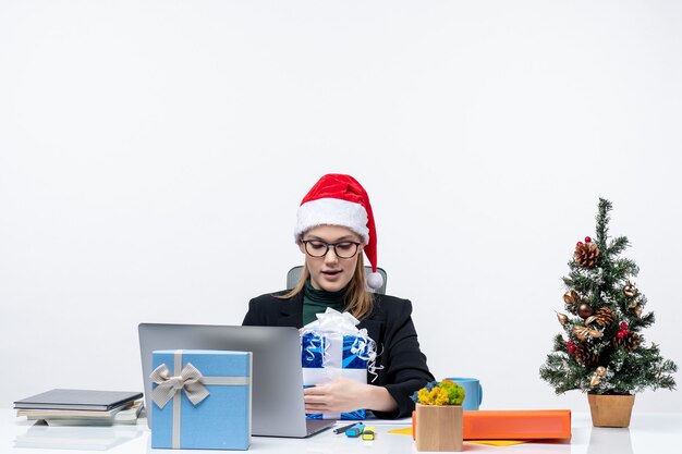 Xsmas mood with happy young woman with santa claus hat and wearing eyeglasses sitting at a table looking at her gift proudly on white background