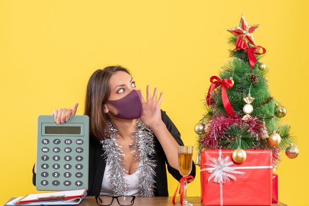 Xsmas mood with serious beautiful lady in suit wearing medical mask holding calculator in the office on yellow 