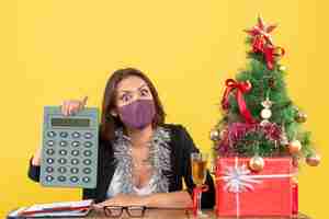 Бесплатное фото Рождественское настроение с потрясенной красивой дамой в костюме в медицинской маске, держащей калькулятор в офисе на желтом