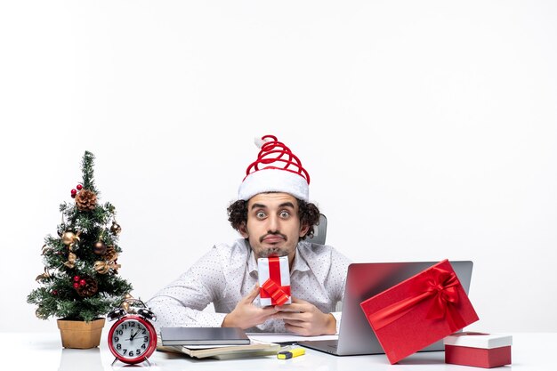 Рождественское настроение с удивленным занятым молодым бизнесменом в шляпе санта-клауса, сидящим в офисе и держащим свой подарок, позирующим перед камерой на белом фоне