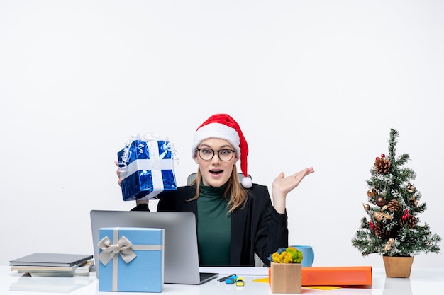 Xsmas mood with surprises young woman with santa claus hat and wearing eyeglasses sitting at a table holding her gift on white background