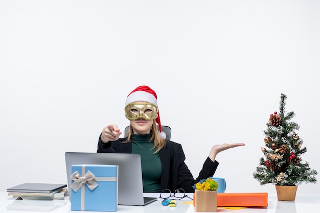 Бесплатное фото Рождественское настроение с напряженной молодой женщиной в шляпе санта-клауса и в маске, сидящей за столом и спрашивающей о чем-то на белом фоне