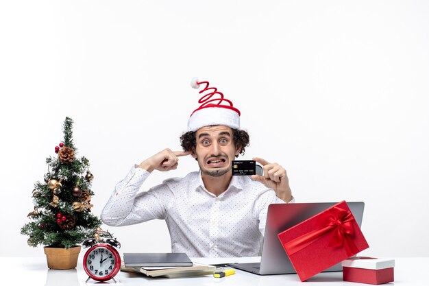 Рождественское настроение с молодым бородатым сердитым деловым человеком в шляпе санта-клауса, держащим банковскую карту и закрывающим ухо в офисе