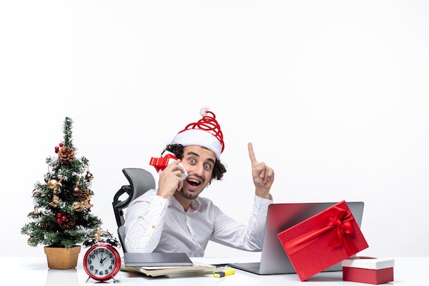 Рождественское настроение с молодым бородатым деловым человеком в шляпе санта-клауса, поднимающим свой подарок и указывающим выше на белом фоне