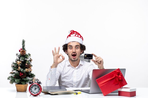 Xsmas mood with young bearded shocked proud business person with santa claus hat holding bank card making eyeglasses gesture on white background