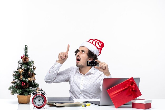 Рождественское настроение с молодым бородатым удивленным деловым человеком в шляпе санта-клауса, держащим банковскую карту и указывающим выше в офисе