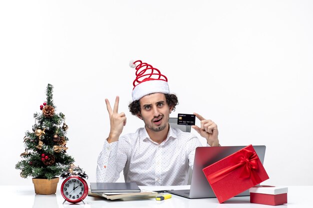 Рождественское настроение с молодым бородатым удивленным деловым человеком в шляпе санта-клауса, держащей банковскую карту и показывающую двоих на белом фоне