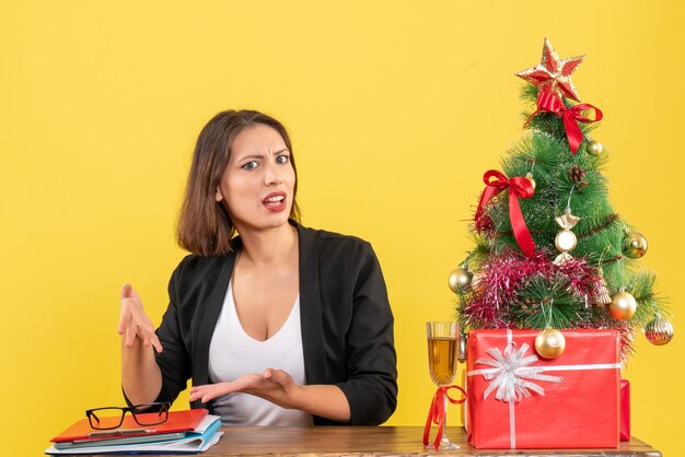 Рождественское настроение с молодой сбитой с толку эмоциональной бизнес-леди, которая что-то спрашивает, сидя за столом в офисе