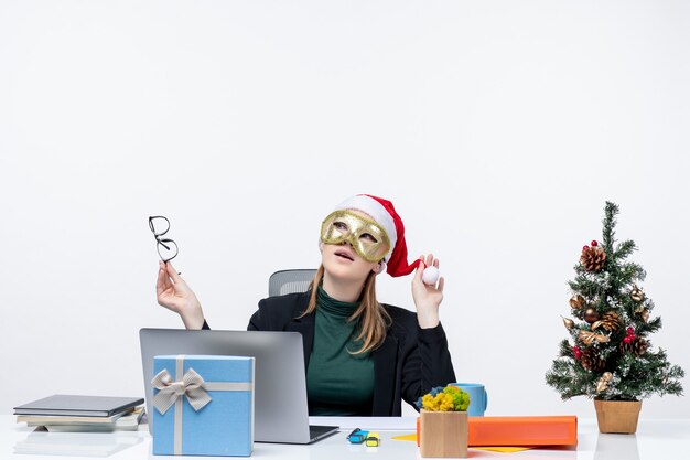 Рождественское настроение с молодой женщиной, играющей в шляпе санта-клауса с очками и в маске, сидящей за столом на белом фоне
