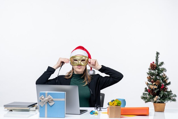 Рождественское настроение с молодой женщиной в шляпе санта-клауса и в маске, сидя за столом на белом фоне stock photo