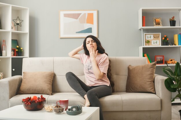 Yawning covered mouth with hand young girl sitting on sofa behind coffee table in living room