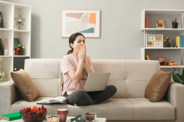 Yawning covered mouth with hands young girl sitting on sofa behind coffee table holding and used laptop in living room