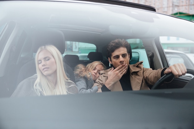 Зевающий человек сидит в машине со спящей женой и дочерью