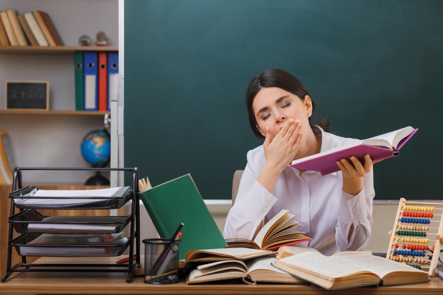 目を閉じてあくび教室で学校の道具を持って机に座って本を持っている若い女性教師