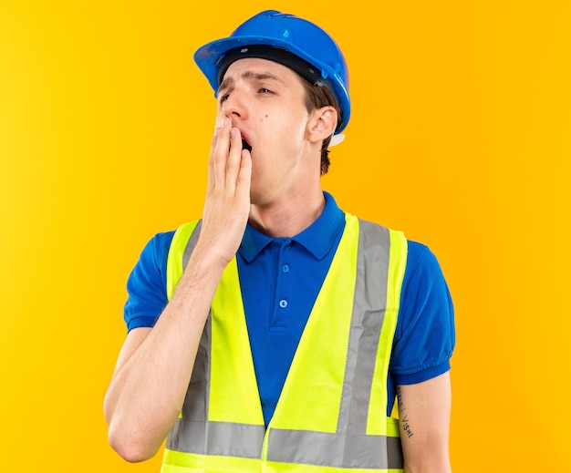 Yawning young builder man in uniform covered mouth with hand isolated on yellow wall